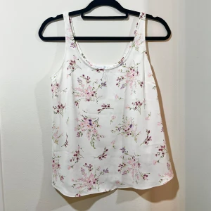 Vit blommig topp  - Oanvänd söt, vit topp i silkesmaterial med blommigt mönster i rosa och lila nyanser. Toppen är ärmlös och har en bekväm passform. Passar bra till jeans eller kjol för en fräsch look.