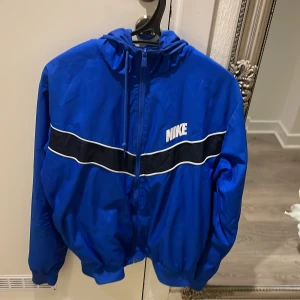 Blå vindjacka från Nike - Säljer en blå vindjacka från Nike i mycket bra skick. Jackan har en dragkedja framtill och en vit Nike-logga på bröstet. Den har en sportig look med svarta och vita ränder över bröstet. Perfekt för vår och höst!
