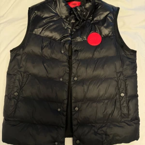 Svart dunväst från Hugo - Säljer en snygg svart dunväst från Hugo i mycket bra skick. Västen har en quiltad design med knappar och en stor röd logga på bröstet. Perfekt för höst och vår när du vill hålla dig varm men ändå se stilren ut. Passar både till vardags och fest!