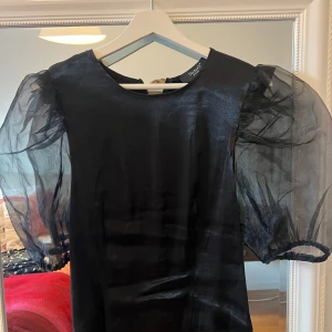 Svart blus med puffärmar - Snygg svart blus från SisterS med puffiga, transparenta ärmar. Perfekt för festliga tillfällen! Blusen har en elegant och stilren design med en dragkedja i ryggen för enkel påklädning.