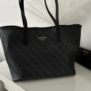Svart axelväska från Guess - Snygg svart axelväska från Guess i mycket bra skick. Väskan har ett diskret mönster och är tillverkad i syntetmaterial. Köpt hos Zalando för 1495kr, den är slutsåld. Den har även svarta fläckar i insidan. Har inte försökt att ta bort men det kanske går bort.