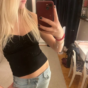 Svart linne med detaljer - Säljer ett svart linne med smala axelband och snygga paljetter framtill. Perfekt för en utekväll eller sommarens fester. Linnet är tight och sitter bekvämt på kroppen. Passar bra till jeans eller kjol för en stilren look.