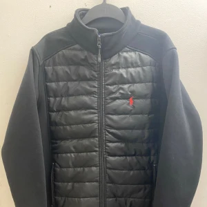 Svart jacka från Ralph Lauren - Säljer en snygg svart jacka från Ralph Lauren i mycket bra skick. Den har en quiltad framsida med dragkedja och det klassiska röda logotypbroderiet. Perfekt för höst och vår, med långa ärmar och en stilren look. Passar både till vardags och lite finare tillfällen. Den har använts jätte sällsynt för att den passar inte mig