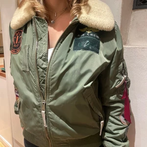 Grön bomberjacka från Alpha Industries - Säljer en grön bomberjacka från Alpha Industries i bra skick. Den har en mysig pälsliknande krage och coola patches på ärmen och bröstet. Jackan har dragkedja och ribbstickade muddar vid ärmslut och nederkant. Perfekt för vintern!