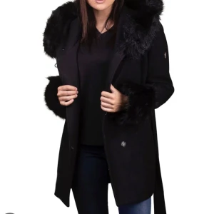 Svart kappa med fuskpälsdetaljer - Säljer en snygg svart kappa med fuskpäls på huva och ärmslut. Perfekt för kyliga höst- och vinterdagar. Den stängs med knappar. Kappan har också ett bälte i midjan för en fin siluett. Supermysig och stilren!