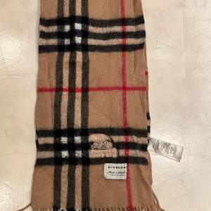 Säljer en klassisk rutig halsduk från Burberry i 100% kashmir. Den har en beige bas med svarta, vita och röda rutor. Perfekt för att hålla sig varm och stilren under kyliga dagar. Halsduken är i mycket bra skick och har fransar i ändarna. Passar perfekt till höst- och vinteroutfits! Pris går  att diskutera!