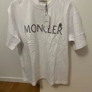 Vit t-shirt från Moncler - Säljer en snygg vit t-shirt från Moncler i nytt skick. Den har en stor Moncler-logga på framsidan och en liten logga på ärmen. Perfekt för en stilren och avslappnad look. T-shirten är kortärmad och gjord i bomull, vilket gör den skön att bära. Kommer med originalförpackning och etikett. Storlek S men sitter lite större…