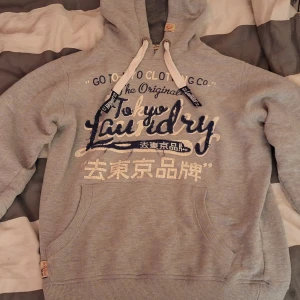 Grå hoodie från Tokyo Laundry - Säljer en grå hoodie från Tokyo Laundry i bra skick. Den har gått sönder vid snöret lite grann, men är ändå snygg med lite vintage stil. Den har en stor, mysig huva med vita dragsnören och en känguruficka framtill. Trycket på bröstet är i marinblått och vitt, vilket ger en cool kontrast. Perfekt för höst och vinter!