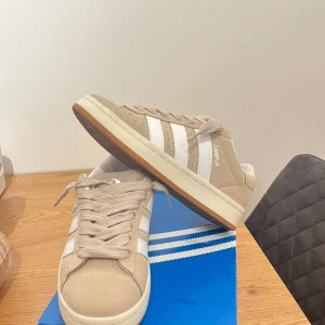 Adidas Campus skor - Säljer ett par snygga beige Adidas Campus sneakers i nytt skick. Dem är endast använda ungefär 4-6 gånger har rengört dom ordentligt. De har klassiska vita ränder och en bekväm gummisula. Perfekta för både vardag och fest! Skorna är i storlek dam 38. Pris kan diskuteras. Dem är ljusa som bild 1 Inte som på bild 2