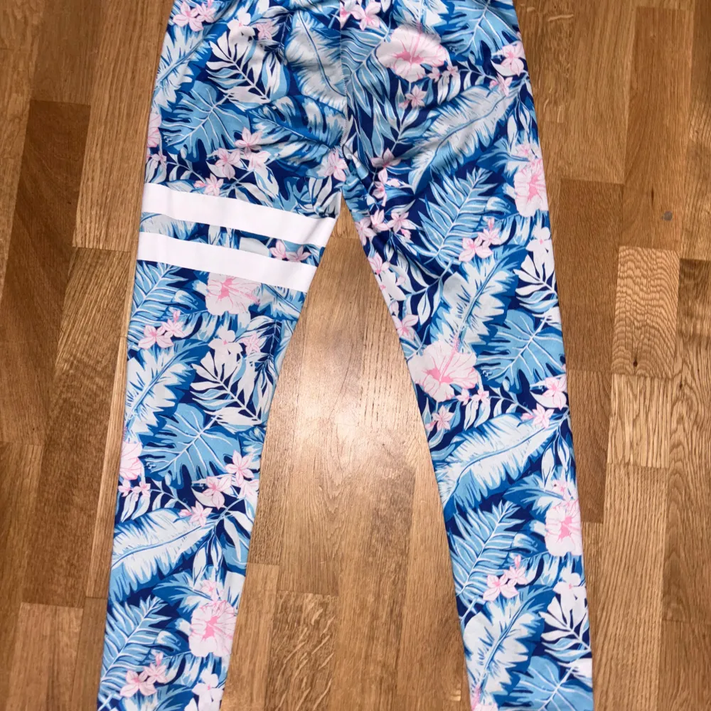 Säljer ett par snygga leggings med ett tropiskt blommönster i blått och rosa. De har vita ränder på ena benet och är perfekta för sommarens alla äventyr. Superbekväma och stretchiga!. Jeans & Byxor.