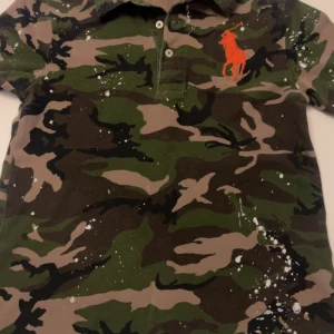 Camouflage pikétröja från Polo Ralph Lauren - Säljer en snygg camouflage pikétröja från Polo Ralph Lauren. Den har en klassisk krage och knappar framtill. Tröjan är i gröna och bruna toner med ett coolt orange logotypbroderi på bröstet. Perfekt för en avslappnad stil! Skick på tröjan är 5/10 skick och tröjan är 8år och small i barn!