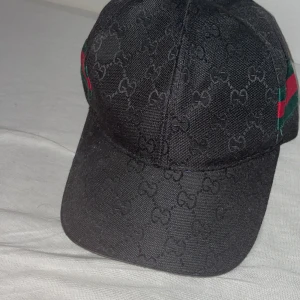 Svart keps med mönster - Snygg svart keps från Gucci med deras ikoniska mönster. Kepsen har en klassisk design med justerbar rem baktill och de typiska gröna och röda ränderna på sidan. Perfekt för att ge din outfit en lyxig touch!