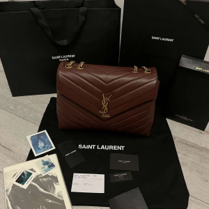 Brun axelväska från Saint Laurent - Nu säljer min syster sin superfina vinröda väska från Saint Laurent . Väskan har ett quiltat mönster och guldfärgade kedjedetaljer. Den är perfekt för både vardag och fest. Väskan är i mycket bra skick och kommer med originalförpackning.
