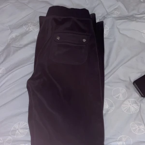  Juicy Couture - Säljer ett par svarta mjukisbyxor från Juicy Couture i storlek S. Byxorna har en bekväm passform med snörning i midjan och fickor med knappar bak. Perfekta för en avslappnad stil eller mysiga hemmakvällar.