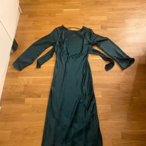 Mörkgrön satin långklänning från Gina Tricot - Säljer en elegant mörkgrön satin långklänning från Gina Tricot. Klänningen har långa ärmar och en stilren design med en öppen rygg. Perfekt för höst- och vinterfester. Materialet känns mjukt och lyxigt, vilket gör den både bekväm och snygg. Passar perfekt för en kväll ute eller en speciell tillställning.