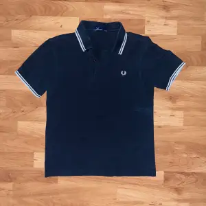 Säljer en snygg svart pikétröja från Fred Perry i storlek S. Tröjan är i bomull och har vita detaljer på kragen och ärmsluten. Perfekt för en stilren och avslappnad look. Den har en broderad logga på bröstet. Passar både till vardags och fest!