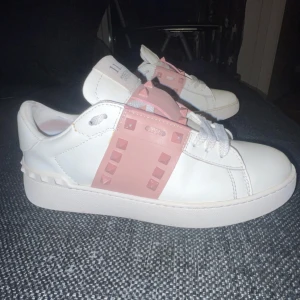 Vita sneakers med rosa detaljer från Valentino - Säljer ett par snygga vita sneakers från Valentino med coola rosa rockstud-detaljer. De är i nytt skick och har en stilren design med snörning. Perfekta för både vardag och fest! Storlek dam 38-39. Original pris ≈8000kr. Priset kan diskuteras för att kvitto och box finns inte.