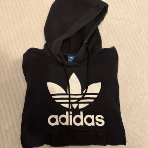 Svart hoodie från Adidas - Säljer en svart hoodie från Adidas med den klassiska loggan i vitt på framsidan. Den är superbekväm och perfekt för både vardag och träning. Hoodien har en justerbar huva och en stor ficka framtill. Passar perfekt till höst och vinter!