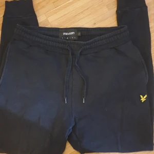 Svarta mjukisbyxor från Lyle & Scott - Säljer ett par svarta mjukisbyxor från Lyle & Scott i bra skick. De har en bekväm passform med resår och snörning i midjan. Byxorna har en liten gul logga på sidan och en bakficka. Perfekta för en avslappnad dag hemma eller en snabb tur till affären.