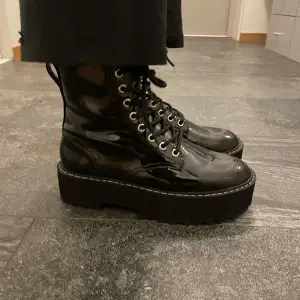 Säljer ett par svarta, glansiga boots i lack med snörning framtill. De har en rejäl platåsula som ger extra höjd och stil. Perfekta för att ge din outfit en edgy touch. Användbara både till vardags och fest!