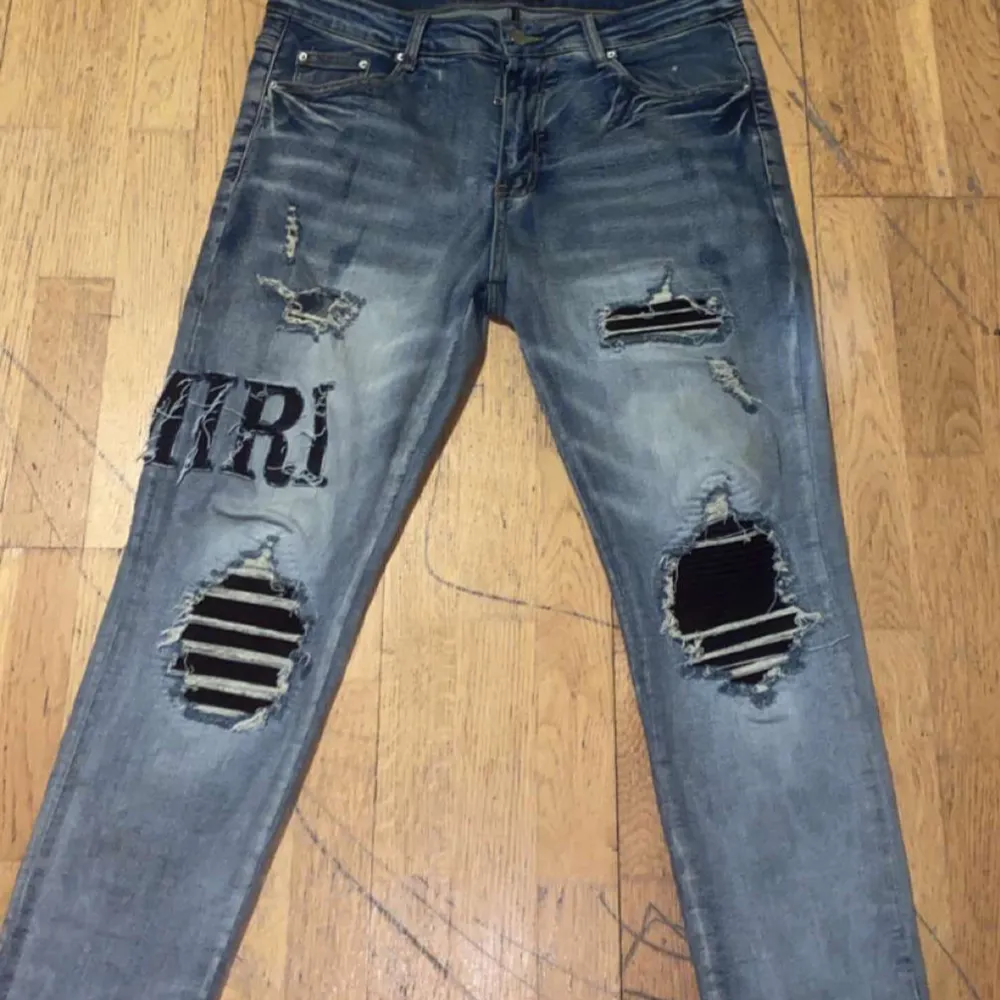 Säljer ett par snygga blå jeans från Amiri med coola slitningar och broderad textdetalj. De har en slim passform och är perfekta för en avslappnad stil. Jeansen har knappar och dragkedja framtill. Perfekta för både vardag och fest!. Jeans & Byxor.