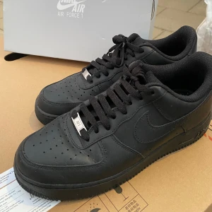 Svarta Nike Air Force 1 '07 - Säljer ett par klassiska svarta Nike Air Force 1 '07 i storlek 42.5. Skorna är i nytt skick och har en stilren design med snörning och perforerade detaljer för extra ventilation. Köpta för 1 395kr. Perfekta för både vardag och fest!