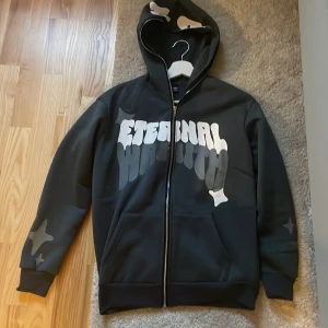 Svart hoodie med tryck - Säljer en svart hoodie med coolt 'Eternal' tryck på framsidan. Den har en dragkedja och ribbade muddar vid ärmsluten. Perfekt för höst och vinter, och den har en skön passform. Huvan har ett mönster som matchar trycket. Perfekt för en avslappnad stil!