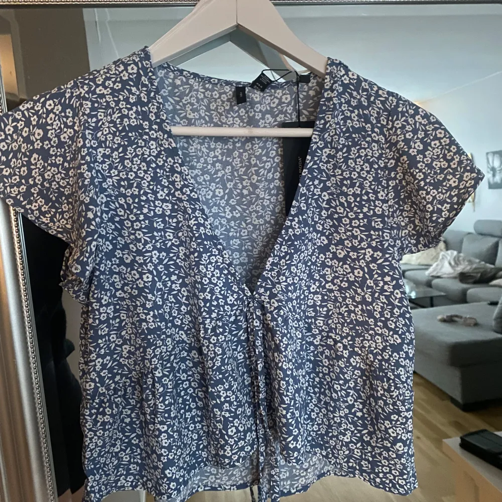 Söt blå blommig blus med korta ärmar och v-ringning. Den har en knytning framtill som ger en fin detalj 🌸. Yläosat.