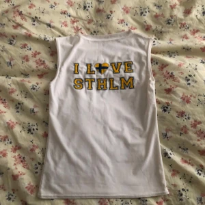 Vit ärmlös topp med 'I Love STHLM' tryck - Säljer en vit tanktop med texten 'I Love STHLM' i blått och gult. Toppen är gjord av 92% polyester och 8% elastan, vilket gör den stretchig. Storlek är uppskattat XS pga ingen lapp. Tillverkad i England.