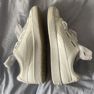 Vita sneakers från Puma med breda snören - Säljer ett par vita Puma sneakers i nyskick. De har en klassisk design med breda, vita snören som ger en unik touch. Skorna är tillverkade i skinn och har en bekväm passform. Perfekta för både vardag och fest! Finns andra skosnören till som du får med!! Dem är i stor storlek 