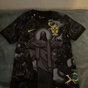 Säljer en svart Brasilien fotbollströja från Nike med ett unikt mönster som inkluderar Kristusstatyn och andra detaljer. Tröjan har korta ärmar och det ikoniska Brasilien-emblemet i gult på bröstet. Perfekt för fotbollsfans som vill ha något speciellt!