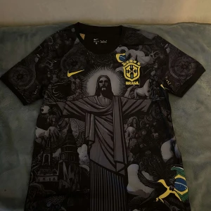 Svart Brasilien fotbollströja från Nike - Säljer en svart Brasilien fotbollströja från Nike med ett unikt mönster som inkluderar Kristusstatyn och andra detaljer. Tröjan har korta ärmar och det ikoniska Brasilien-emblemet i gult på bröstet. Perfekt för fotbollsfans som vill ha något speciellt!