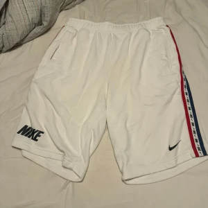 Vita shorts från Nike - Säljer ett par vita Nike shorts i storlek M. De har en elastisk midja och är superbekväma för både träning och vardag. Snygga detaljer med röd och blå rand längs sidorna och Nike-logga på benet. Perfekta för sommaren!