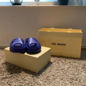 Blå sandaler från Axel Arigato - Säljer ett par snygga blå tofflor från Axel Arigato. De är sprillans nya (aldrig använda)och har en modern design med en solid blå färg. Tofflorna har en bekväm passform och är perfekta för sommaren. Sulan har ett bra grepp för både inne och utomhus.