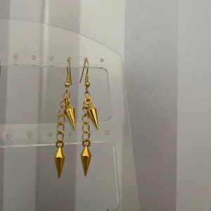 Guldörhängen med kedja och konformad detalj - Säljer ett par snygga guldörhängen med en kedja och en nit längstner. De är perfekta för att ge en edgy touch till din outfit. Passar både till vardags och fest! Egengjorda! 