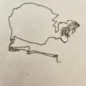 Silverhalsband med bokstavsberlock - Säljer ett stilrent silverhalsband med en liten bokstavsberlock 'L'. Kedjan är tunn och elegant, perfekt för vardagsbruk eller som en personlig present. Halsbandet har en enkel och tidlös design.