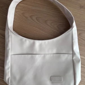 Vit handväska - En helt ny och oanvänd vit väska! Har en ficka på framsidan och jättemycket plats inuti så det rymmer ganska mycket! Den har dessutom bred axelband så det ska vara skönt på axeln. Mått: 38x25x10cm