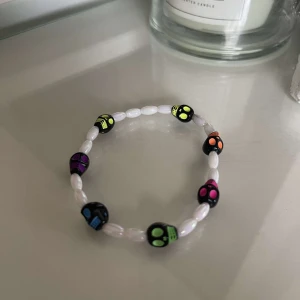 Färgglatt armband med dödskallar - Säljer ett coolt armband med vita pärlor och färgglada dödskallepärlor i olika färger som lila, grön, blå och orange. Perfekt för att ge en edgy touch till din outfit!