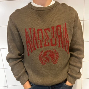 Brun stickad tröja med rött tryck - Säljer en snygg brun stickad tröja med ett coolt rött tryck på framsidan. Tröjan har långa ärmar och en rund halsringning. Perfekt för höst och vinter, och den är superbekväm att ha på sig. Passar både till vardags och lite mer avslappnade tillfällen.