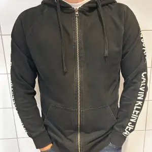 Säljer en snygg svart hoodie från Calvin Klein Jeans. Den har en dragkedja framtill och text längs ärmarna. Perfekt för en casual look eller när du bara vill vara bekväm. Den är i bra skick och redo för en ny ägare!