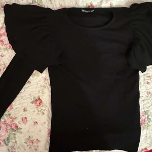 Svart top med volang - Ribbad, svart långärmad tröja med söta volanger på axlarna. 