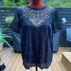 Svart topp med detaljer Lindex 40 - Svart topp med påsydda metalldetaljer Längd: 66 cm Axelbredd: 42 cm Viskos & polyester  Inga bilder när den sitter på mig! Katter finns i hemmet