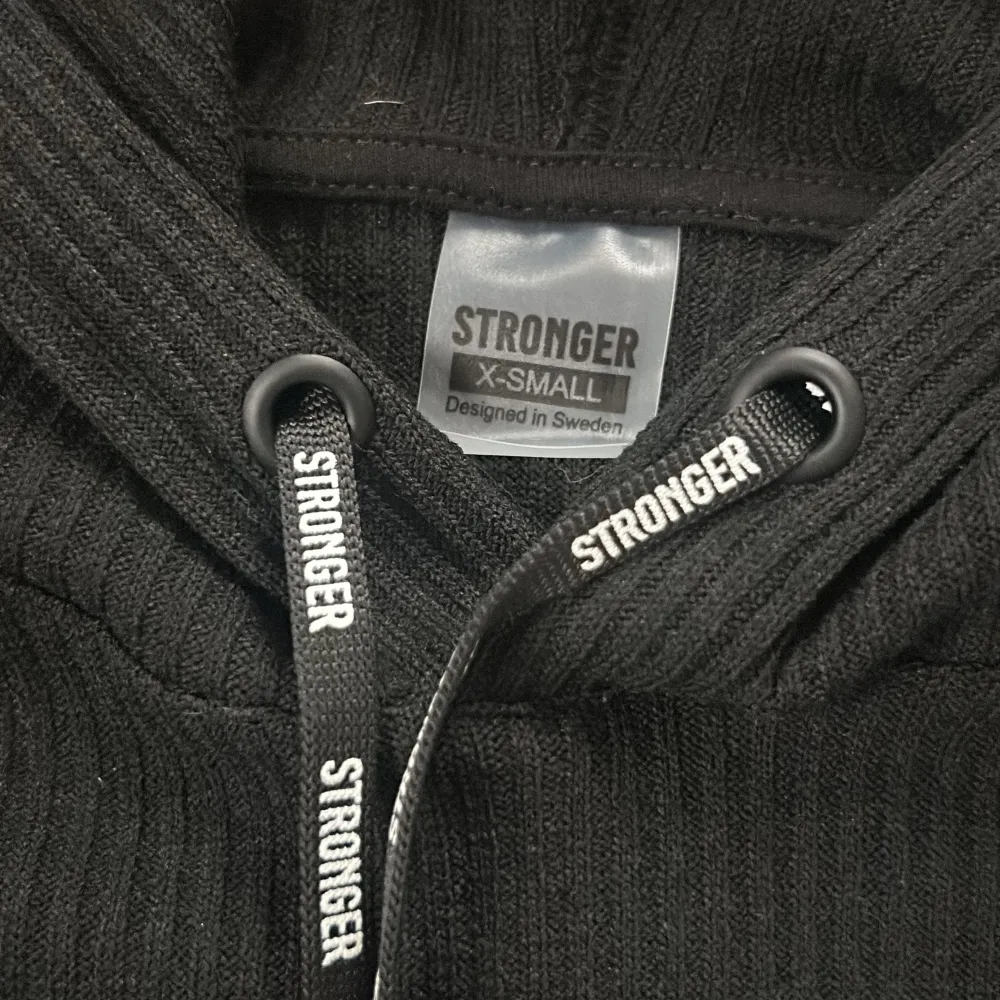 Säljer en snygg svart croppad hoodie från Stronger. Den är i storlek XS och har en cool ribbad design. Perfekt för både träning och vardag! Den har långa ärmar och en justerbar huva med snören där det står 'STRONGER'.. Sportswear.