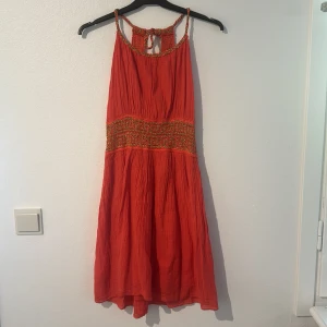 Röd sommarklänning med broderade detaljer - Säljer en superfin Korall sommarklänning med vackra broderade detaljer i guld.  Den är ärmlös och har en fin knytning i ryggen där både längd osv går att justera!❤️