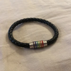 Svart flätat armband med regnbågsfärgad detalj - Säljer ett snyggt svart flätat armband med en cool regnbågsfärgad detalj i mitten. Armbandet har en stilren design med en magnetisk låsning i silverfärg. Perfekt accessoar för både vardag och fest!