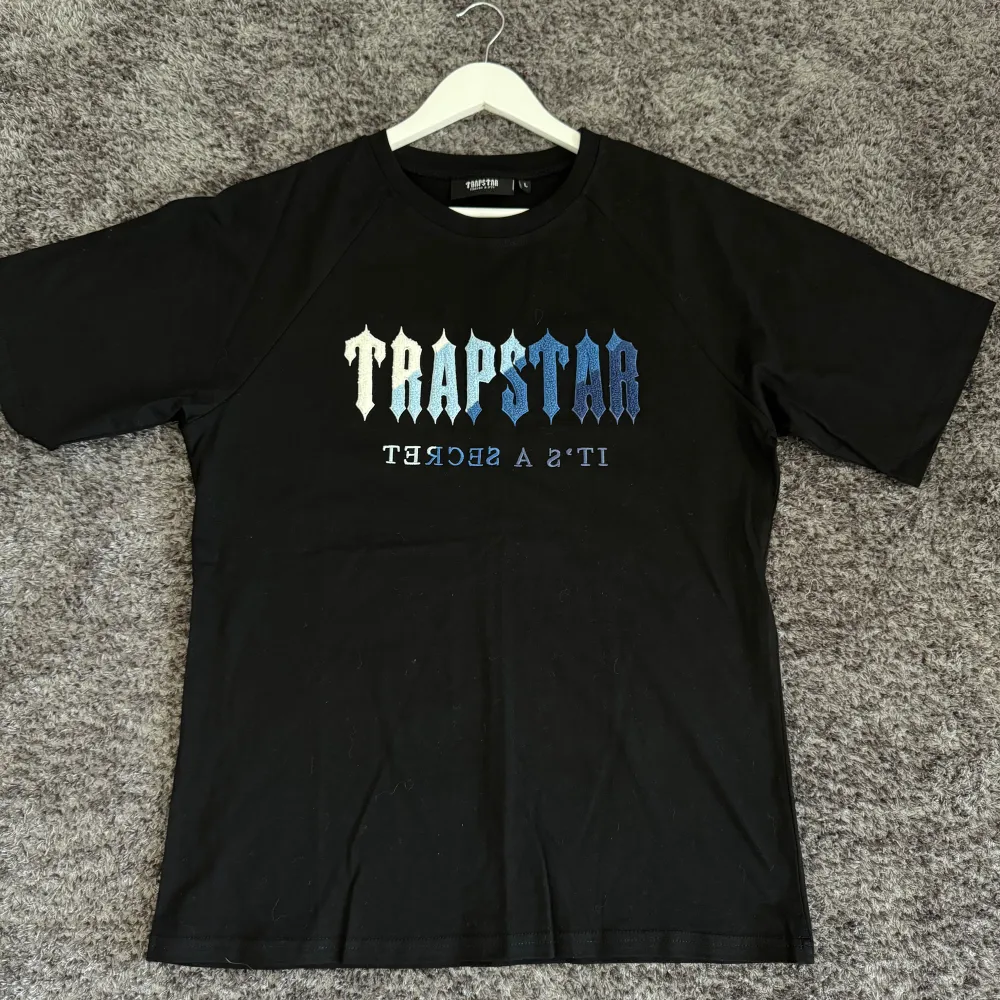 Säljer en svart t-shirt från Trapstar med texten 'IT'S A SECRET' i blå och vit gradient på framsidan. T-shirten är i storlek L och har en klassisk passform med korta ärmar. Perfekt för en casual look!. T-shirts.