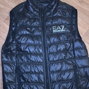 Svart quiltad dunväst från Emporio Armani - Säljer en snygg svart quiltad dunväst från Emporio Armani. Västen har en dragkedja framtill och är perfekt för lager-på-lager under höst och vår. Den är i nyskick och har en stilren design med märkets logga på bröstet.