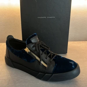 Svarta och blå sneakers från Giuseppe Zanotti - Säljer ett par snygga sneakers från Giuseppe Zanotti i svart och blått. Skorna har en cool design med dragkedjor i guld på sidorna och snörning framtill. De är gjorda i en kombination av skinn och sammet vilket ger dem en lyxig känsla. Äktahetsbevis✅