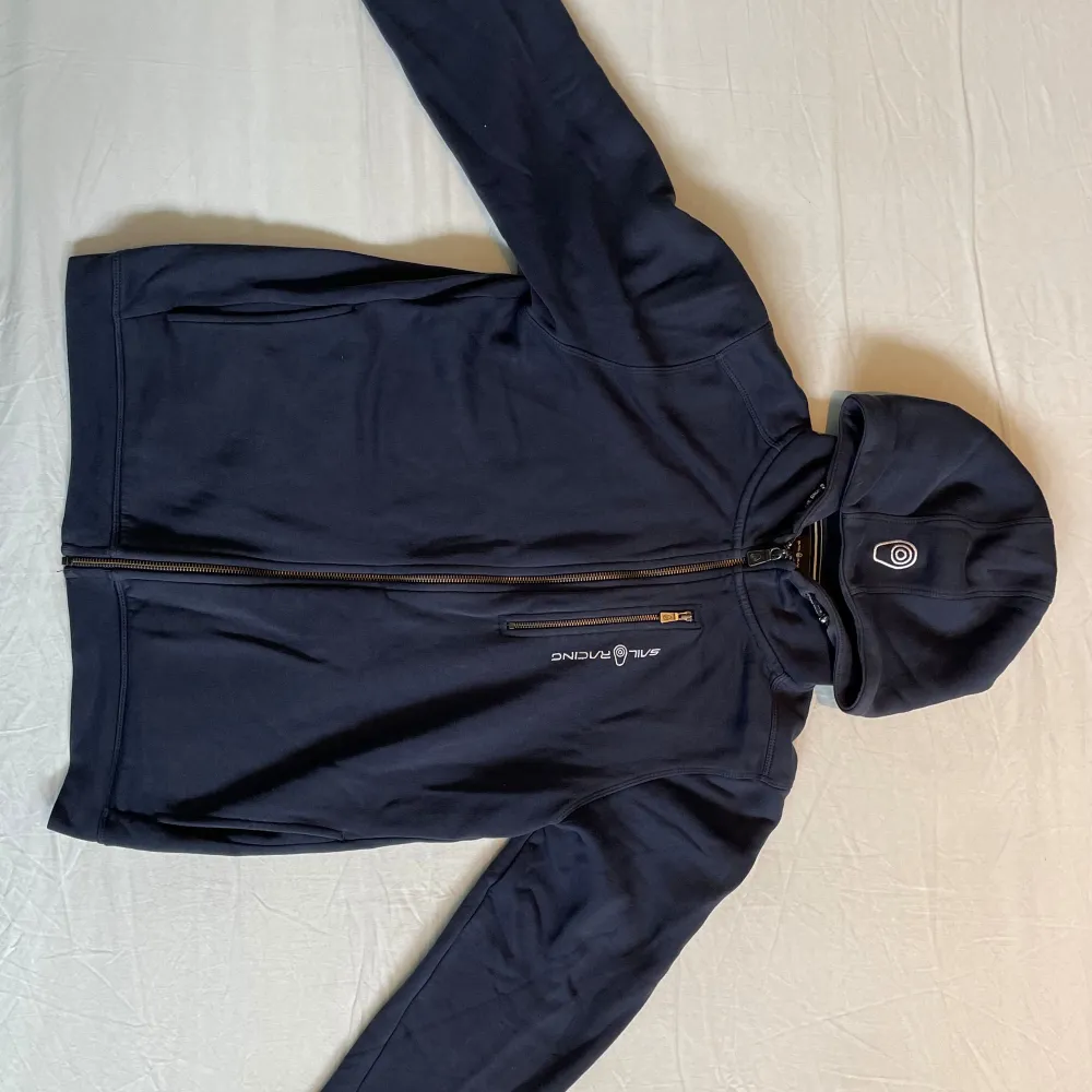 Säljer en mörkblå zip hoodie från Sail Racing i storlek 176 (passar även stl S). Den har en dragkedja framtill och en huva. Insidan är fodrad med mjukt material. Det finns ett litet märke på högra ärmen, men annars i bra skick. Kom gärna med prisförslag och frågor.. Hoodies.