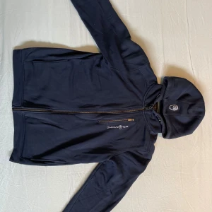 Mörkblå zip hoodie från Sail Racing - Säljer en mörkblå zip hoodie från Sail Racing i storlek 176 (passar även stl S). Den har en dragkedja framtill och en huva. Insidan är fodrad med mjukt material. Det finns ett litet märke på högra ärmen, men annars i bra skick. Kom gärna med prisförslag och frågor.